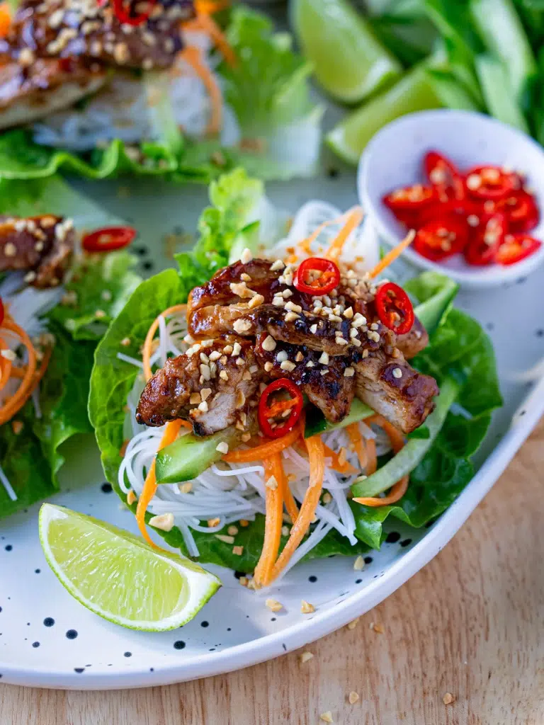 Hoisin chicken served in lettuce cups with vermicelli noodles, julienned carrots, cucumber, crushed peanuts, and sliced red chili, garnished with lime wedges on the side.